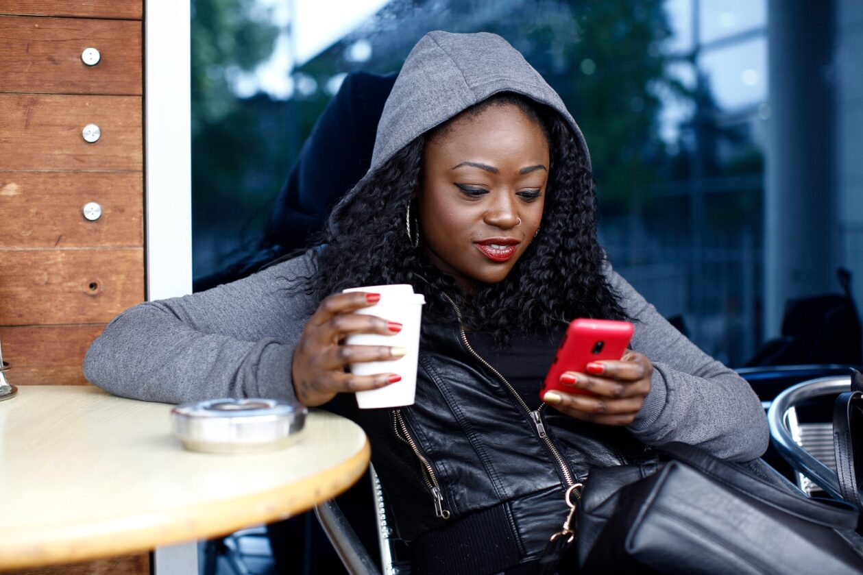 Woman seated outdoors at a round table, using a red cell phone in her left hand and holding a coffee in her right hand.