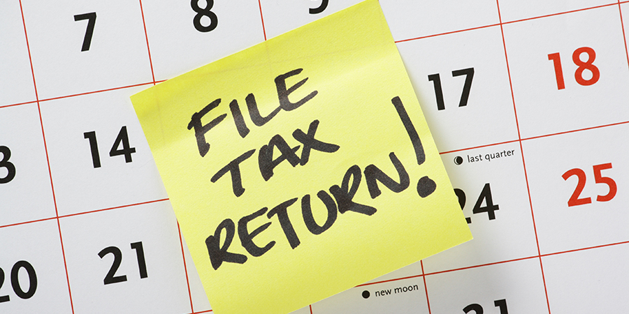 Yellow post-it note with “FILE TAX RETURN!” written in marker placed on a calendar in between the 14th and 17th days.