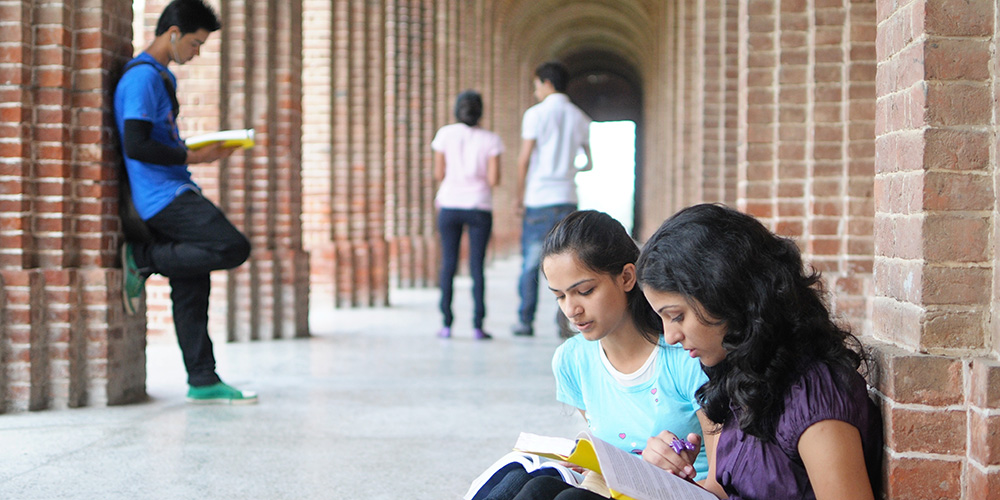 Five students in an outside campus corridor, including two seated by a wall, reading class material funded by the LLC.